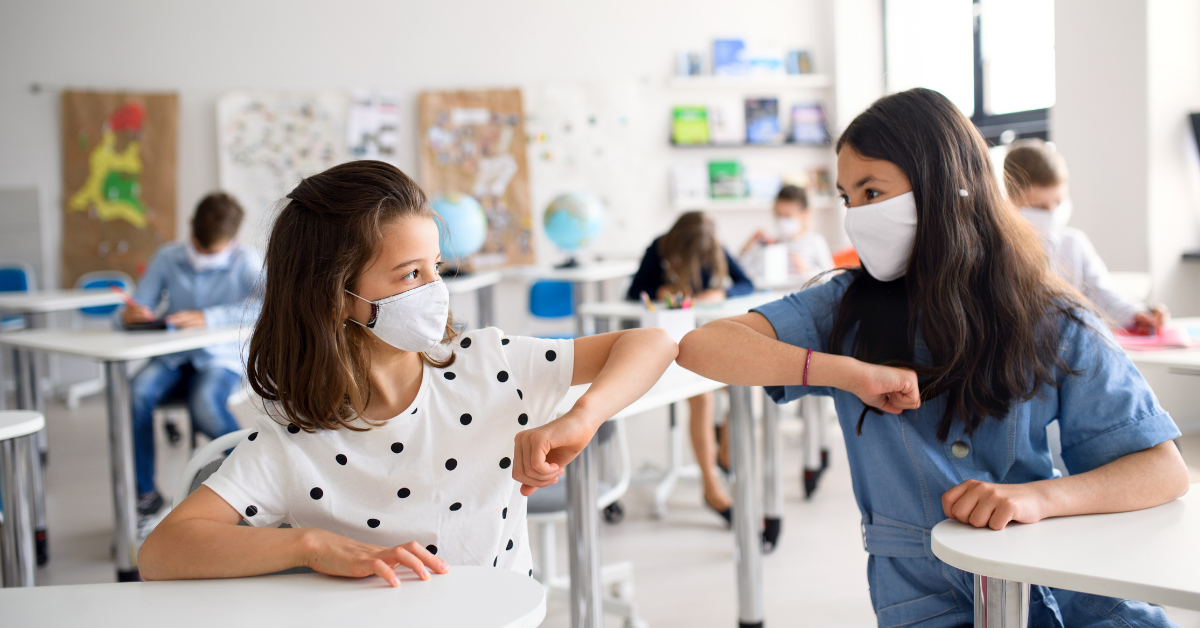 In a school classroom two girls in face masks bump elbows
