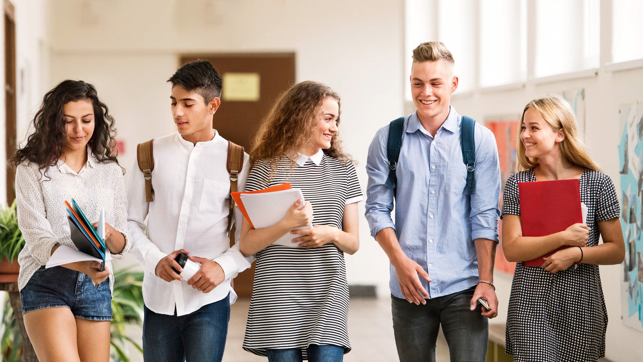 A group of five teens walk shoulder-to-shoulder down a school hallway