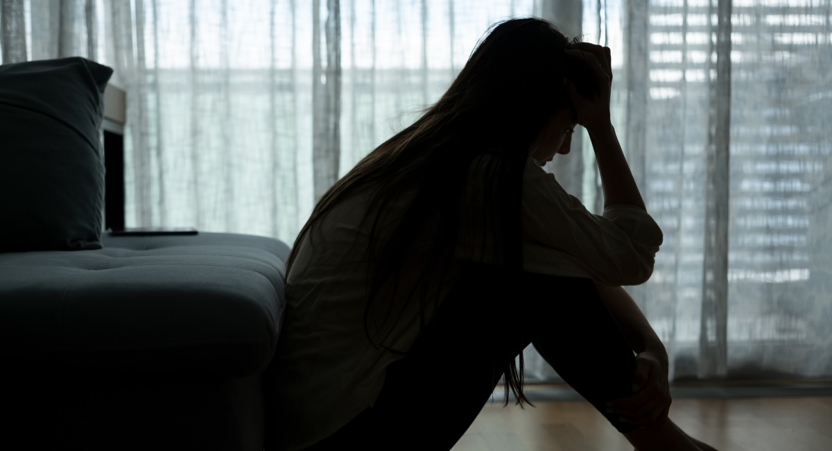 A depressed woman sits alone in her room with her head in her hands in front of a window on a cloudy day