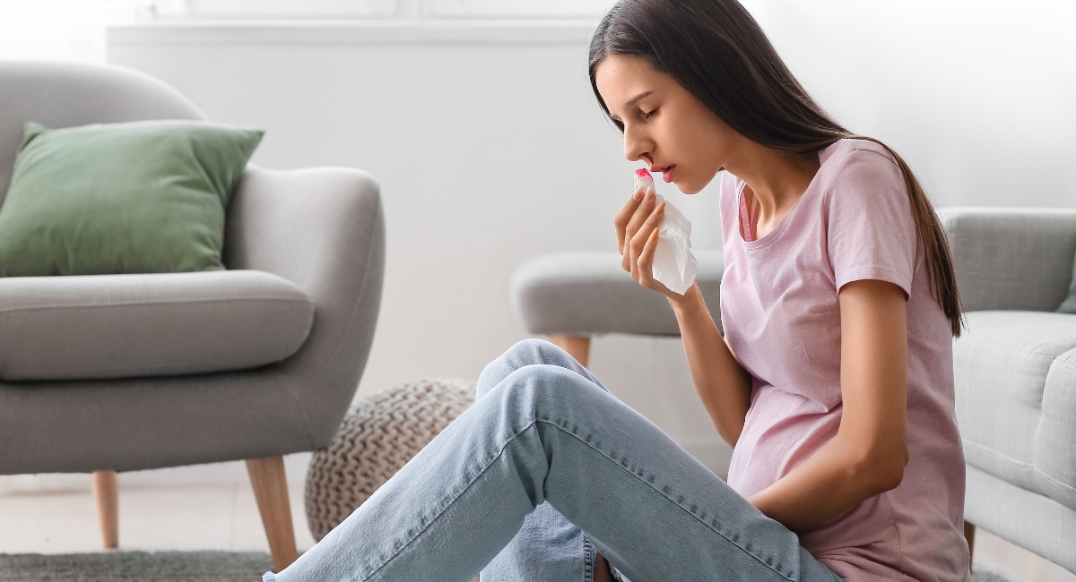 A young woman is seated in on her living room floor, with a tissue under her nose. She is visibly uncomfortable and experiencing a nosebleed.
