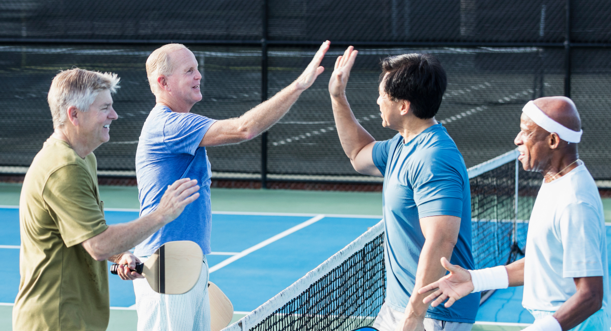 A group four middle aged men exchange handshakes and high fives after a friendly game of pickle ball.