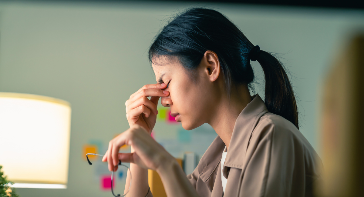 An anxious young woman is seated at her desk. She is holding her eyeglasses in her left hand, and she's using her right hand to find a pressure point near her eyes to relieve a stress-induced headache.