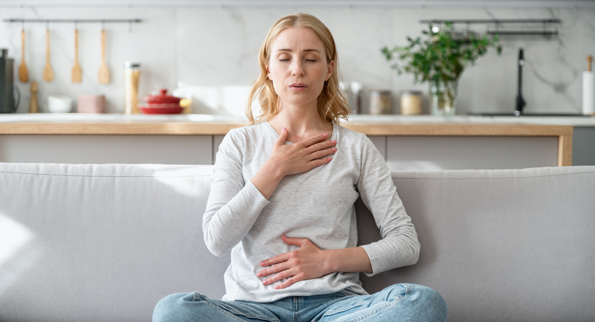 A woman in her 40s is sitting on her couch with her eyes closed. Her right hand is over her heart and her left is on her stomach. She is practicing deep breathing and mindfulness techniques.