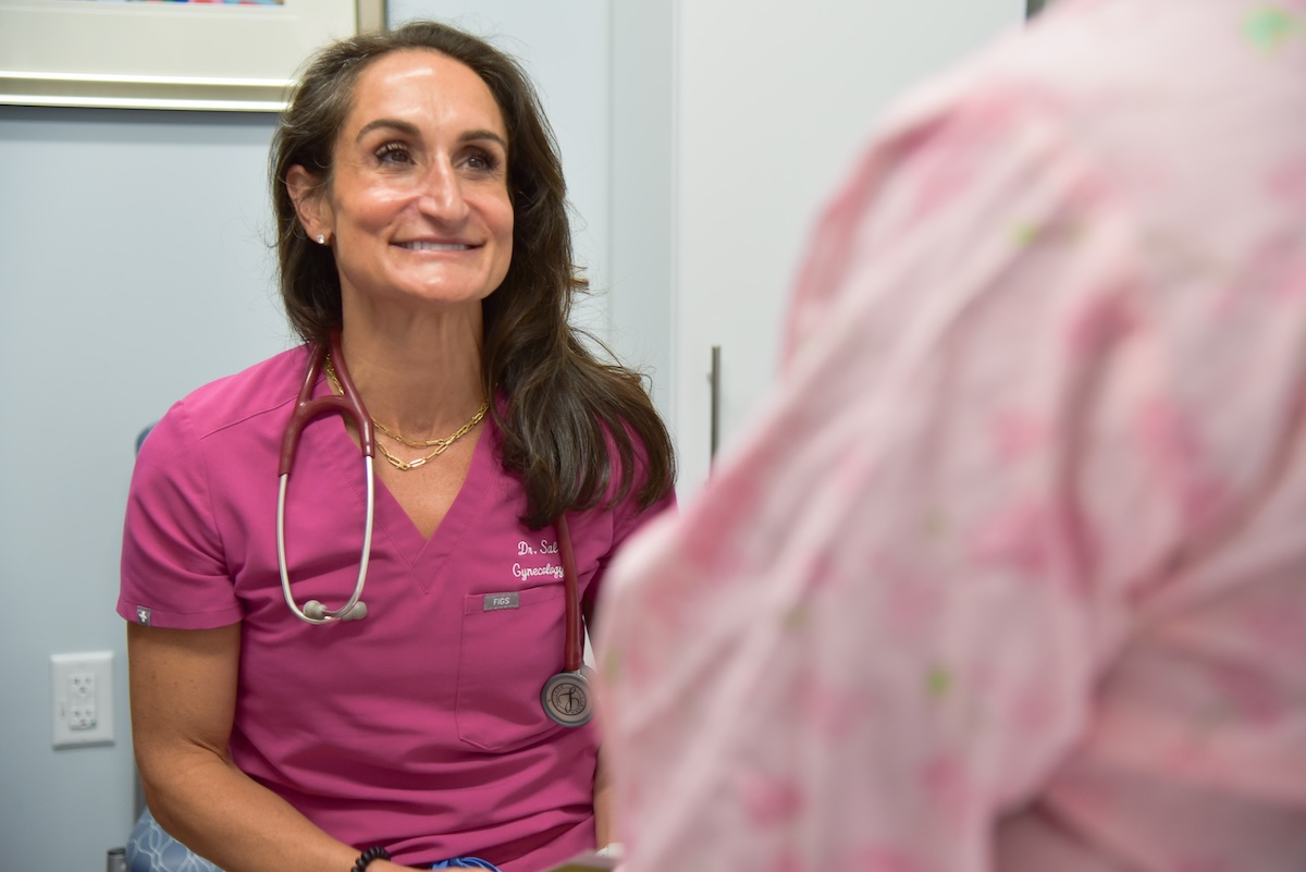 Dr. Salvemini sits in an exam room, talking with a female patient about sexually transmitted infections. She is wearing hot pink scrubs and a stethoscope.