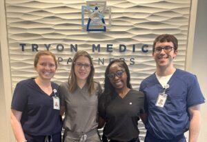 Dermatology medical assistant interns stand in a row from left to right in front of a wall at Tryon Medical Partners. Standing left to right are Sarah Taylor, Madison McBride, Ketsia Kasongo, and Logan Robinson.